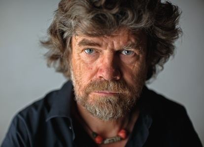 Reinhold Messner seriously looking with a gray short hair, a beard, and a mustache, wearing a necklace and a black polo