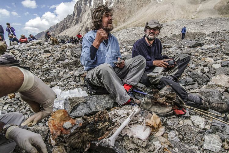 Reinhold Messner (left), brother of Gunther Messner, sitting along with a man, with other mountaineers in their background, they found Gunther Messner’s remains with a femur, clothes, and shoes. Reinhold has a gray short hair with a beard and a mustache, wearing red socks, black shoes, a navy blue long sleeve under a denim jacket, and gray pants, and the man beside him has a beard and mustache, wearing a hat, eyeglasses, black socks, and shoes, navy blue jacket, and gray pants