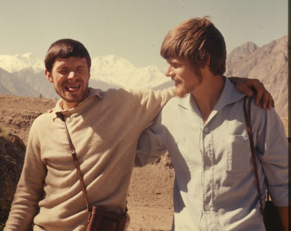 Gunther Messner (left) is smiling with closed eyes while biting his tongue and his right arm around Reinhold Messner's (right) shoulder with a mountain in their background in an old photograph. Gunther has a black hair, a beard, and mustache, wearing a brown shoulder bag and a white polo under a white long sleeve jacket while Reinhold smiling and looking at Gunther while his arms around Gunther’s waist, he has a blonde hair, a beard, and mustache, wearing a brown shoulder bag and a white polo