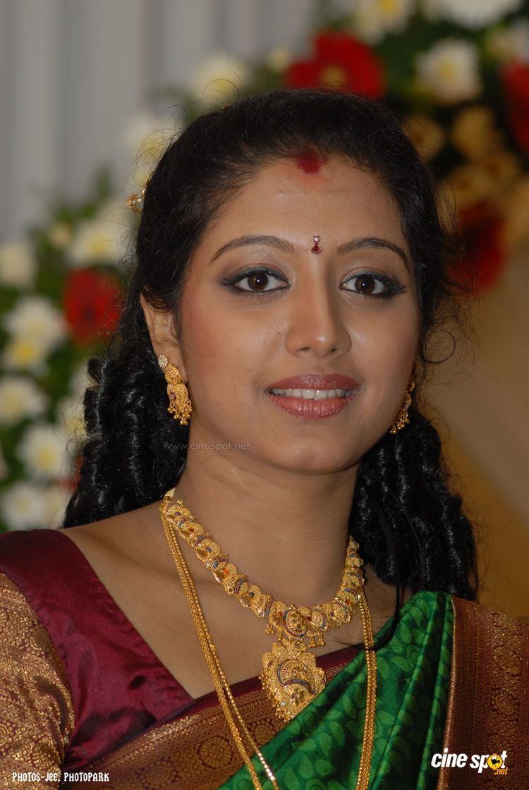 Gopika smiling while wearing a brown, maroon, and green dress, three layered gold necklace, and earrings