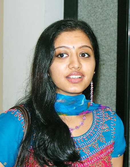 Gopika smiling while looking at something and wearing a blue and pink dress, blue scarf, purple necklace, and earrings