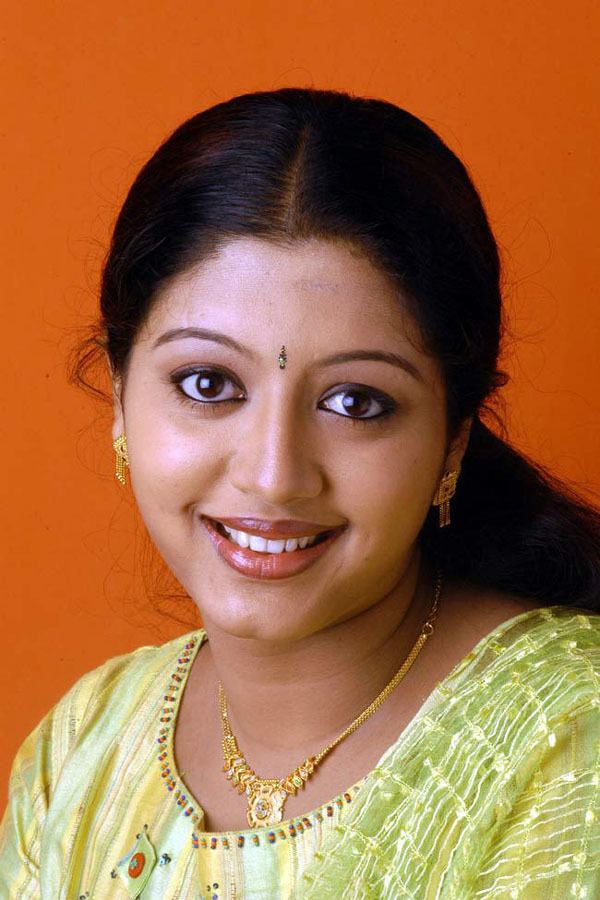 Gopika smiling while wearing a yellow and green dress, yellow dupatta, gold necklace, and earrings