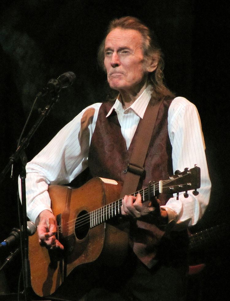 Gordon Lightfoot wearing brown vest and white stripped long sleeves while singing and playing guitar