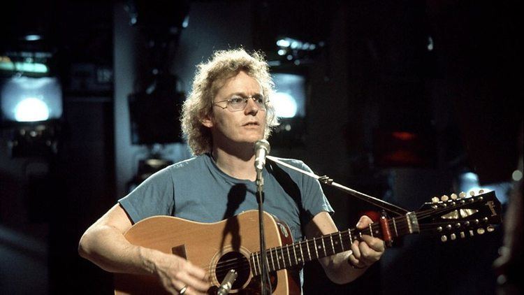 Gordon Lightfoot wearing eyeglasses and gray t-shirt while singing and playing guitar