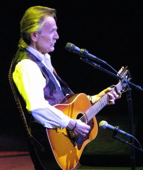 Gordon Lightfoot wearing black vest and white long sleeves while singing and playing guitar