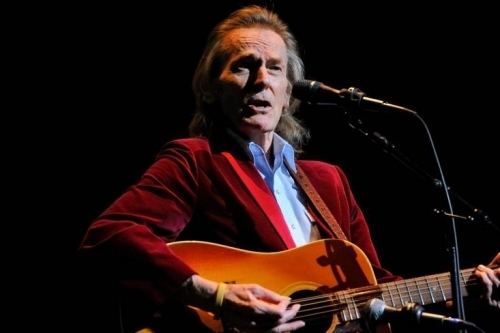 Gordon Lightfoot wearing red coat and blue long sleeves while singing and playing guitar
