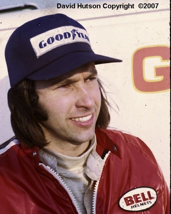 Gordon Smiley smiling while wearing a cap, red jacket and beige turtle neck