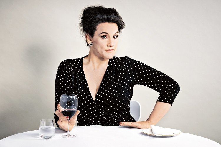Grace Dent holding a glass of wine while the other hand is on her hips and she is wearing a black and white polka dot blouse