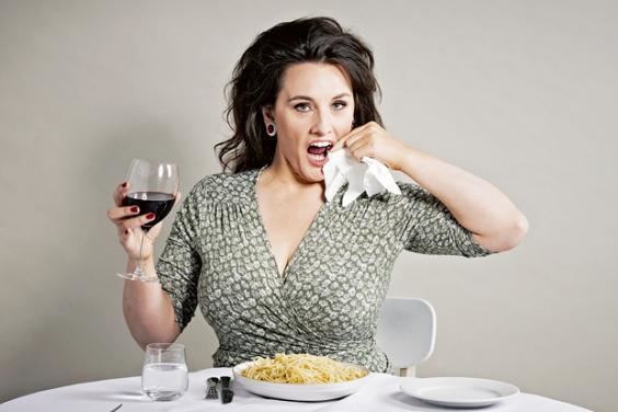 Grace Dent wiping her lips with a tissue, holding a glass of wine, and sitting on the chair while wearing a white and green dress