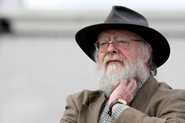 Graham Ovenden with a serious face while holding his neck, with beard and mustache, wearing a black hat, eyeglasses, and brown coat over a checkered long sleeve.