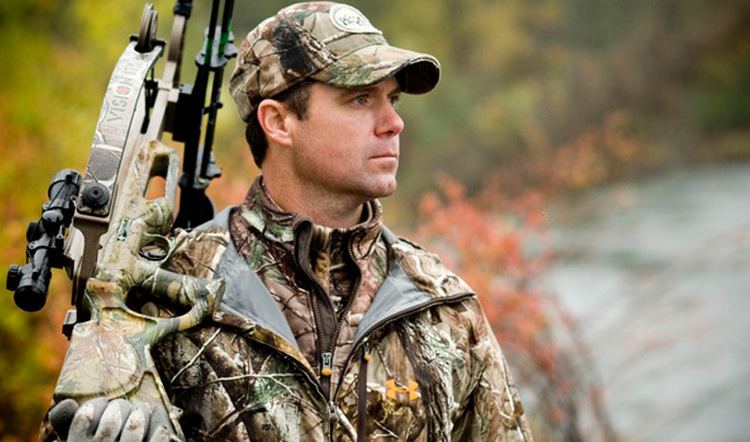 Gregg Ritz looking afar while holding a crossbow and wearing a brown and green cap and jacket