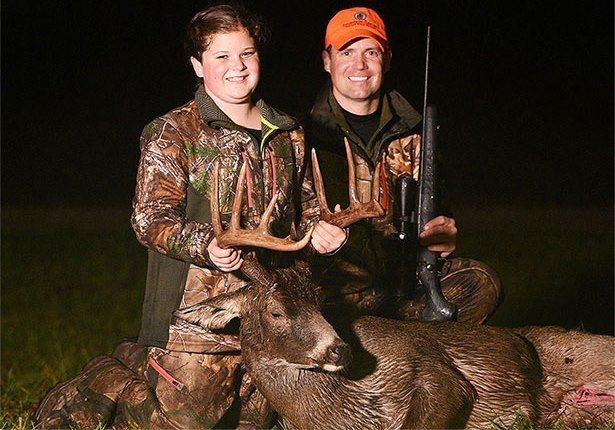 Sienna and Gregg Ritz are smiling while holding the antlers of deer and they are both wearing a brown and green jacket