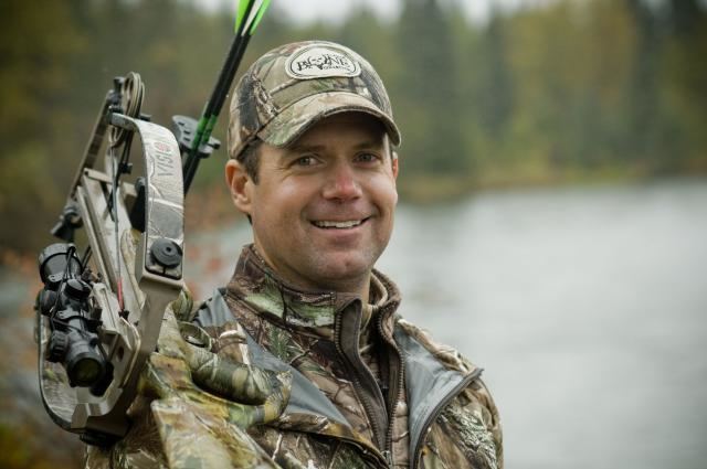 Gregg Ritz smiling while holding a crossbow and wearing a brown and green cap and jacket