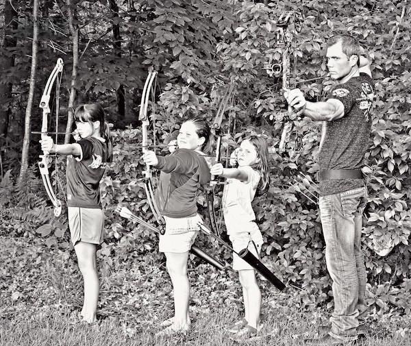 Gregg Ritz and his three daughters are holding arrows and bows while Gregg is wearing a t-shirt and pants and his daughters are wearing t-shirts and shorts