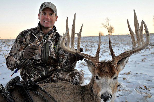 Gregg Ritz smiling and holding an arrow and the antlers of deer while wearing a brown and green cap and jacket