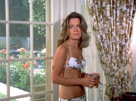 Gretchen Corbett standing and holding a glass of water while wearing a swimsuit