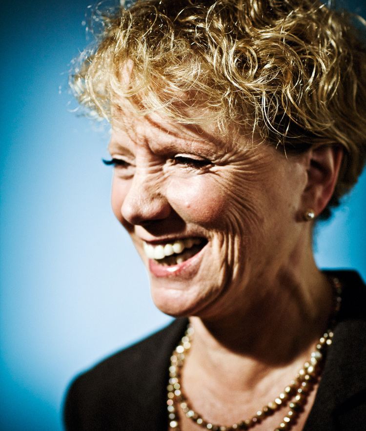 Gretchen Corbett smiles while wearing a necklace and a black sleeve in blue and white background