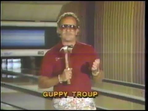 Guppy Troup standing and holding a hammer while wearing shade and a red shirt