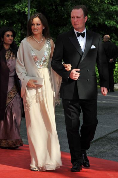 Carina Axelsson (left) is smiling, has black hair, left hand holding the right hand of Gustav, Hereditary Prince of Sayn-Wittgenstein-Berleburg, walking on the red carpet, she wears silver earrings, a necklace, and an off-white sleeveless floral design dress with coat. Behind (on left) is a woman smiling, has black hair, walking on the red carpet, wearing silver earrings, a brown dress with a gold design, and a black coat. Gustav, Hereditary Prince of Sayn-Wittgenstein-Berleburg (right) is serious, has black hair, and his right hand holding the left hand of Carina, walking on the red carpet he wears white long sleeves, a black bow tie, under a black suit, and a pocket on left with a white handkerchief, and black shoes. The man behind (right) is bald, wearing a black suit, pants, and shoes.