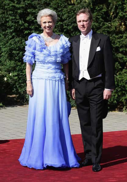 Princess Benedikte (left) is smiling, has white hair, standing on the red carpet, left hand holding a blue purse, wears silver earrings, a necklace, a bracelet on both of her hands, and a  blue long dress. Gustav, Hereditary Prince of Sayn-Wittgenstein-Berleburg (right) is serious, has brown hair, standing on the red carpet, wears white long sleeves, a white bow tie under a black suit, and a pocket on left with a white handkerchief, black pants, and shoes.