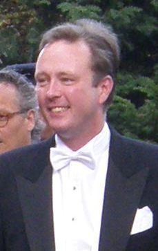 Gustav, Hereditary Prince of Sayn-Wittgenstein-Berleburg is smiling, has brown hair, wearing white long sleeves, a white bow tie under a black suit with a pocket, and a white handkerchief on left. Behind him (on left) is a man, who has white hair, and wearing red eyeglasses.