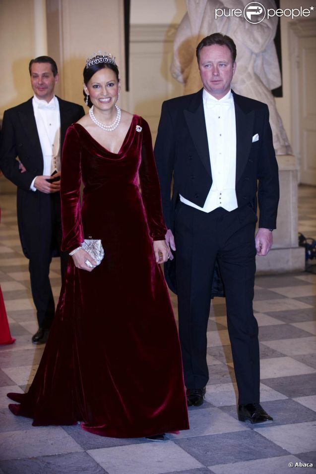 Carina Axelsson is smiling, has black hair, right hand holding a silver purse, she is wearing a silver tiara, silver earrings, a white pearl necklace, a silver bracelet on her right hand, and a red long-sleeve cleavage-showing dress with a pin on left. Behind her is a man smiling, has black hair, both hands holding each other, wearing white long sleeves under a black suit, pants, and black shoes. Gustav, Hereditary Prince of Sayn-Wittgenstein-Berleburg is serious, has black hair, wears a wristwatch on his left hand, and white long sleeves with black buttons under a black suit with a pocket and a white handkerchief, black pants, and shoes. Behind him on left is a white statue.