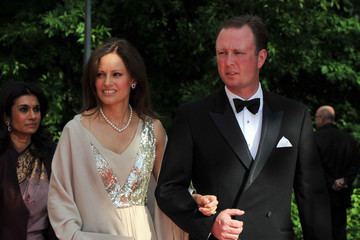 Carina Axelsson (left) is smiling, has black hair, left hand holding the right hand of Gustav, Hereditary Prince of Sayn-Wittgenstein-Berleburg, she wears silver earrings, a necklace, and an off-white sleeveless floral design dress with coat. Behind (on left) is a woman smiling, has black hair, and wears silver earrings, a brown dress with a gold design, and a black coat. Gustav, Hereditary Prince of Sayn-Wittgenstein-Berleburg (right) is serious, has black hair, right hand holding the left hand of Carina, he wears white long sleeves, a black bow tie, under a black suit, and a pocket on left with a white handkerchief. The man behind (right) is bald, wearing a black suit and pants.