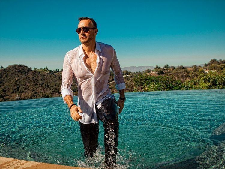 Gyorgy Gattyan with a serious face while standing beside an infinity pool, wearing sunglasses, white wet long sleeves, and wet black pants.