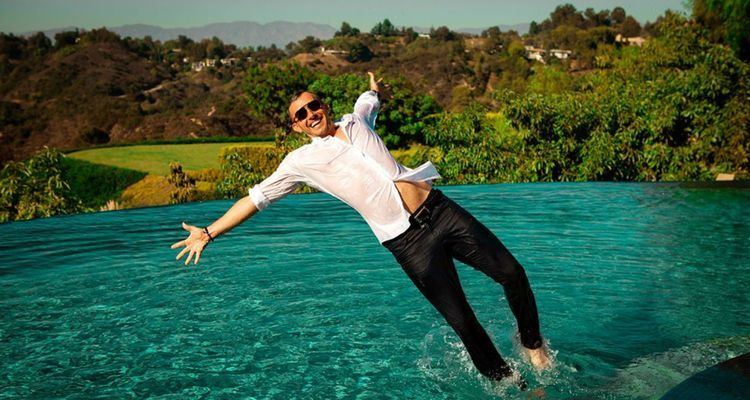 Gyorgy Gattyan smiling while doing a back dive into an infinity pool, wearing sunglasses, white wet long sleeves, and wet black pants.