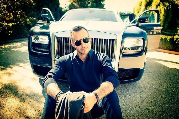 Gyorgy Gattyan with a serious face while sitting on the ground in front of a Black Rolls-Royce Phantom car, wearing sunglasses, blue long sleeves, and blue pants.
