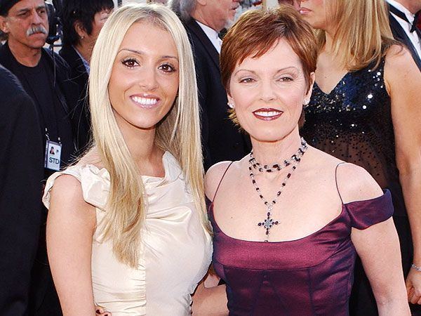 Haley Giraldo smiling with her blonde hair down and wearing a white dress with her mom Pet Benatar with short hair, wearing a maroon dress and a necklace with a cross pendant
