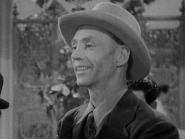 Hank Worden smiling while wearing a hat, coat, long sleeves, and necktie