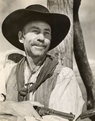 Hank Worden smiling while holding a gun and wearing a hat, long sleeves and vest