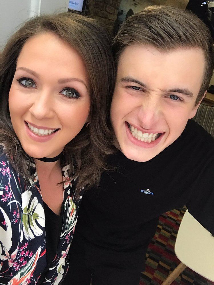 Hannah Hobley smiling while wearing a blue floral blouse and black choker while the man beside him wearing a black t-shirt