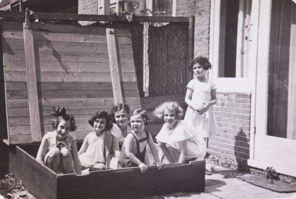 Hanneli Goslar, Anne Frank, Dolly Citroen, Hannah Toby, Barbara Ledermann, and Susanne Ledermann (from left to right), 1937, smiling while sitting inside a big box except for Susanne Ledermann who is standing on the right side and they are all wearing a dress
