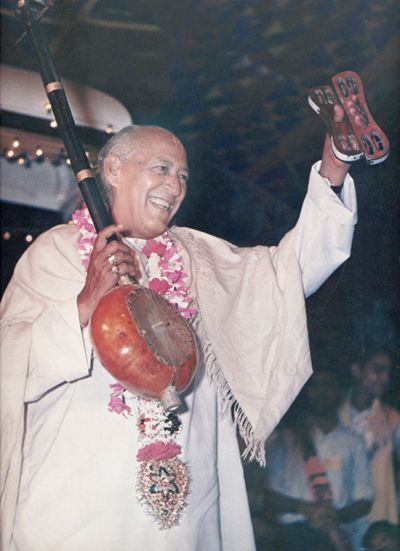 Hans Ji Maharaj smiling while holding a banjo, wearing a flower garland, and off-white garments.