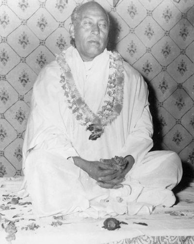Hans Ji Maharaj with closed eyes while sitting on the floor, wearing a flower garland, and white garments.
