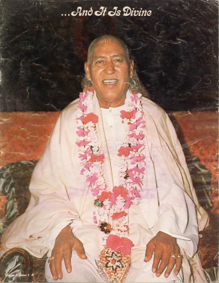 Hans Ji Maharaj smiling while sitting on a couch, wearing a flower garland, and white garments.