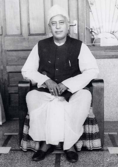 Hans Ji Maharaj with a tight-lipped smile while sitting on a chair, wearing a white Taqiyah, black vest, white garments, and black shoes.