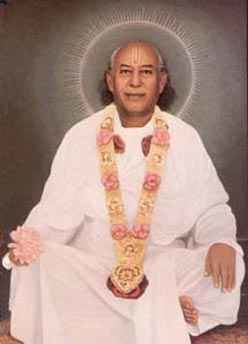 Hans Ji Maharaj smiling while sitting on the floor, wearing a flower garland, and white garments.