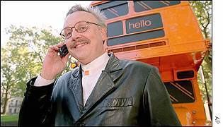 Hans Snook is smiling while holding a mobile phone in front of an orange vehicle. Hans with white hair, wearing eyeglasses and a black leather jacket over a white shirt.