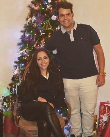 Hariel Ferrari smiling and sitting in front of a Christmas Tree  wearing a black blouse and leggings together with her husband
