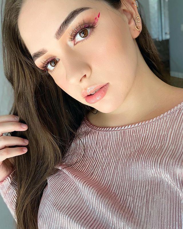 Hariel Ferrari looking serious with her hair down and wearing thick and curly eyelashes, pink eyebrows, earrings and pink and blouse