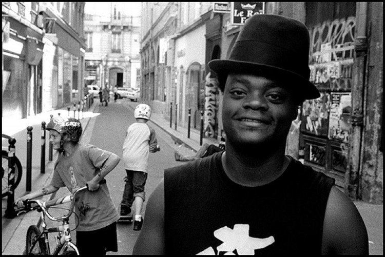 Harold Hunter smiling and wearing a sleeveless shirt and hat while two boys are behind him holding a bicycle