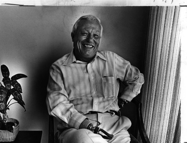 Harold Russell smiling and sitting on a chair while wearing a long sleeve with a plant beside him