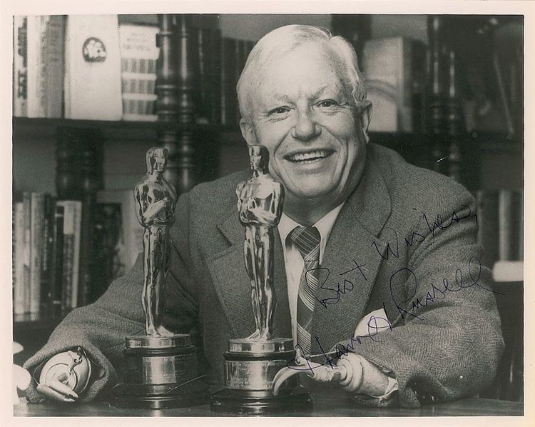 Harold Russell sitting and smiling while wearing a suit with two trophies in front of him