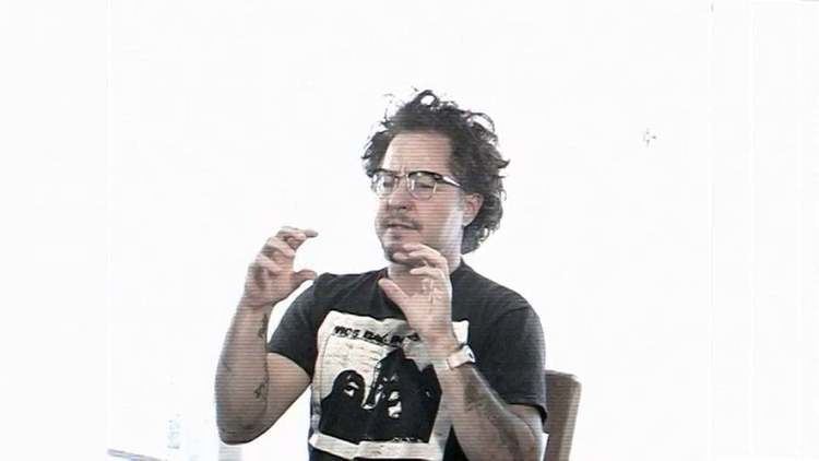 Harry Dodge sitting on the chair and showing hand gestures while wearing a printed t-shirt, wristwatch, and eyeglasses
