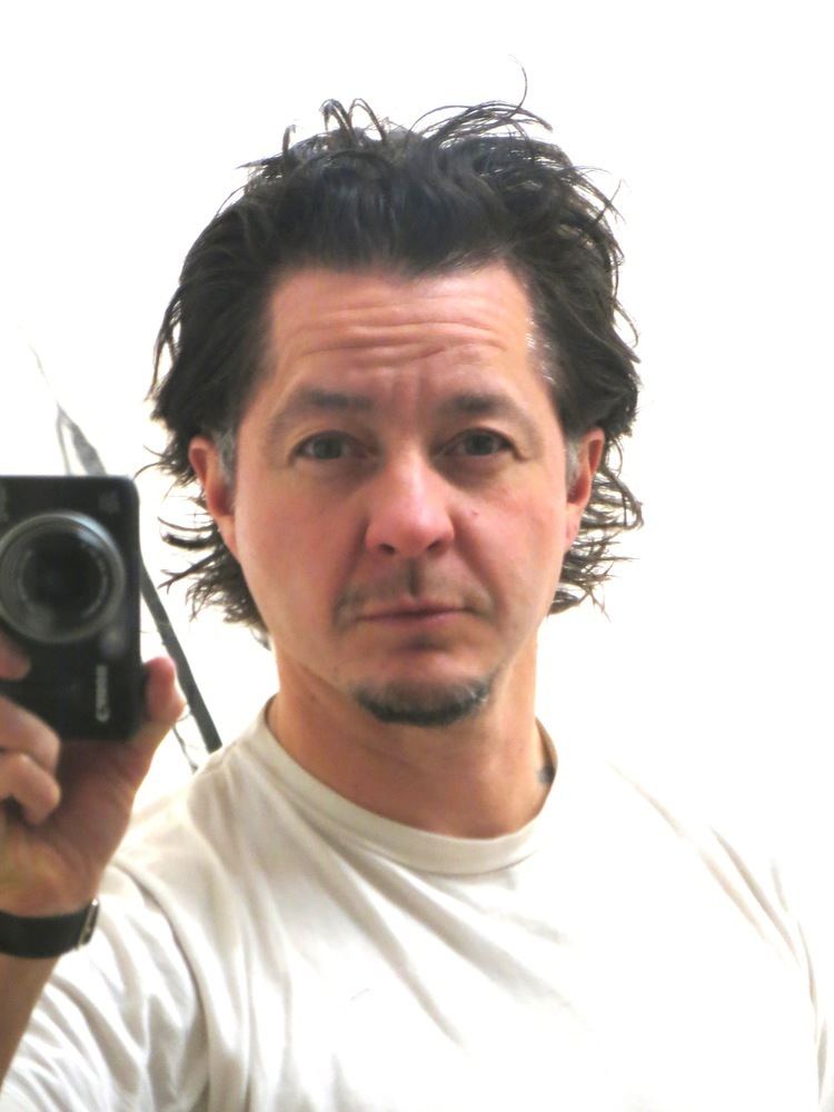 Harry Dodge holding the camera, with a mustache, beard, and a furrowed forehead while wearing a white t-shirt and wristwatch