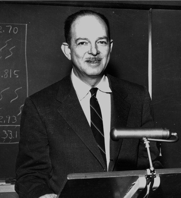 Harry Hess smiling with a mustache, a blackboard behind him, and a tribune podium in front of him while he is wearing a long sleeve under a striped necktie and coat