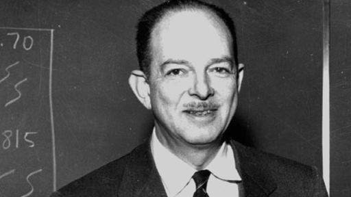 Harry Hess smiling with a mustache and a blackboard behind him while he is wearing a long sleeve under a striped necktie and coat